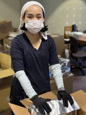 A woman worker with facial mask, head scarf and in gloves putting a sealed package of tea into a carton