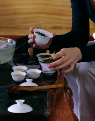 A visiting guest holding a gaiwan in each hand, both filled with tealeaves. Another gaiwan also filled with leaves in the background on the infusion table, together with small cups of tea.