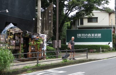 Leo dragging a suitcase case, stopping by a streetside vendor next to a sign directing traffic to the Hakone Open Air Museum