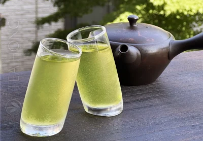 two glasses of cold green tea, frosted with tiny water droplets on the glass wall outside and holding clear light green tea inside, with a kyūsu Japanese teapot in the background, similarly frosted