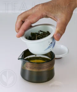 A hand lifting a porcelain infuser from a glass teapot, with a drop of tea dripping from the infuser into the teapot.