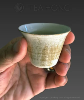 a hand using the thumb and three fingers holding a traditional small teacup with a relatively wider opening that sharply curves into the narrower body