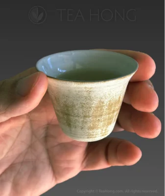 a hand using the thumb and the index finger holding only the widest part of the top of a traditional small teacup with a relatively wider opening that sharply curves into the narrower body