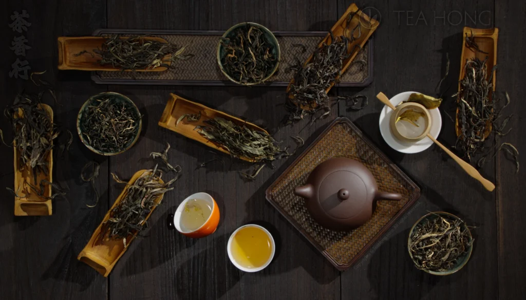 Different kinds of maocha, in various shapes, sizes and colours in bamboo tea scoops or other tea utensils, around a zisha teapot and a few cups of tea