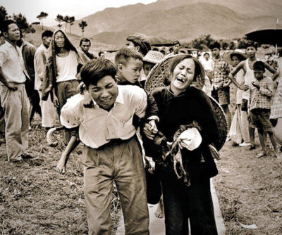 A man carrying a child on the back, and a woman following in dramatically tragic expression, walking away from a crowd of onlookers