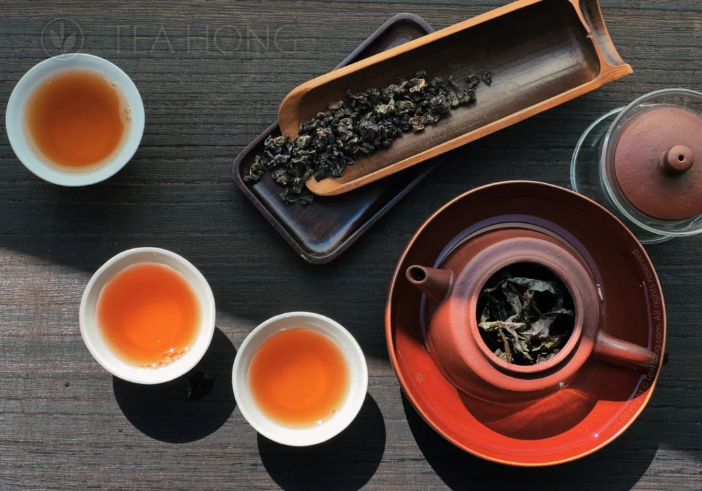 A Yixing teapot with the lid open to show the infused tealeaves of Muzha Tieguanyin in side. Next to it are three small cups, each filled with the intensely amber colour, yet very clear tea liquor.