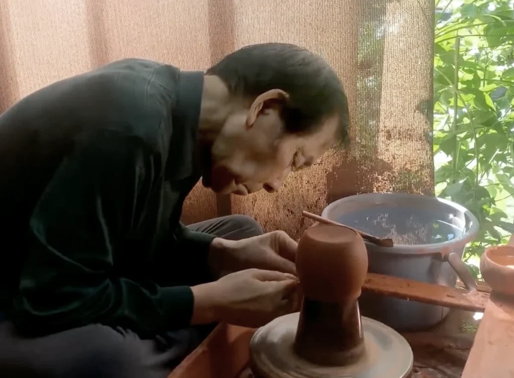 An old artist working at a detail on a red clay pot body over a turning wheel next to a sunlit window, screened by the dense foliage outside