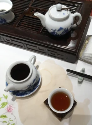 a porcelain teapot on a small infusion stand on a restaurant table covered in white tablecloth with embroidered flowers yet stained with spilled tea. A cup holding the poor looking liquor of Pu'er tea on the side.