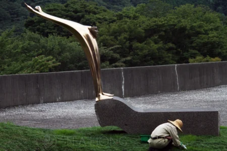 A worker attending to the lawn under and shinny bronze sculpture of a man in a shocked expression, raising two arms